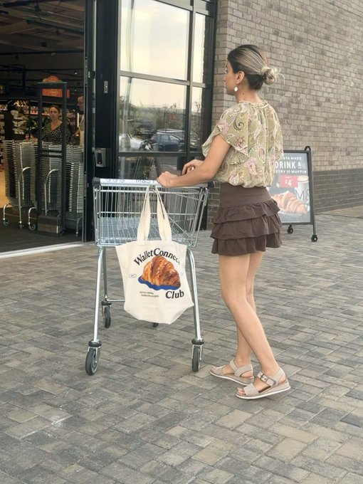 A woman with blonde hair in a bun, wearing a green floral top and brown ruffled skirt, pushing a shopping cart outside a Whole Foods store with a 'WalletConnoisseur Club' tote bag hanging from it.