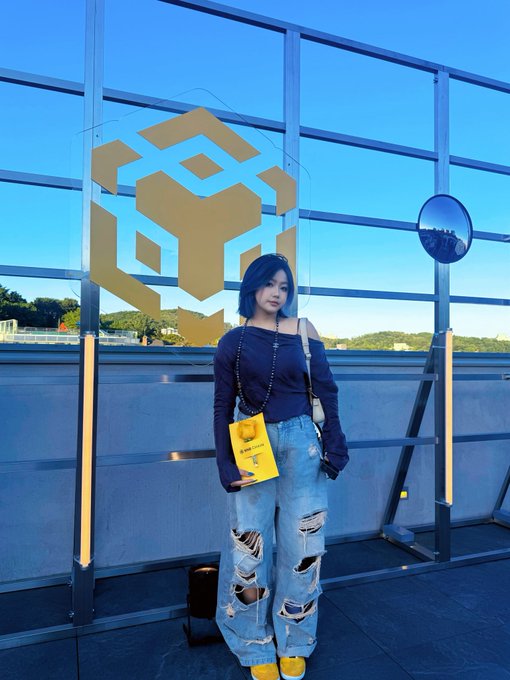 A person with blue hair wearing a dark top and ripped jeans poses in front of a glass wall displaying the BNB (Binance Coin) logo, holding a yellow LEGO bag with a BrickLive ticket.