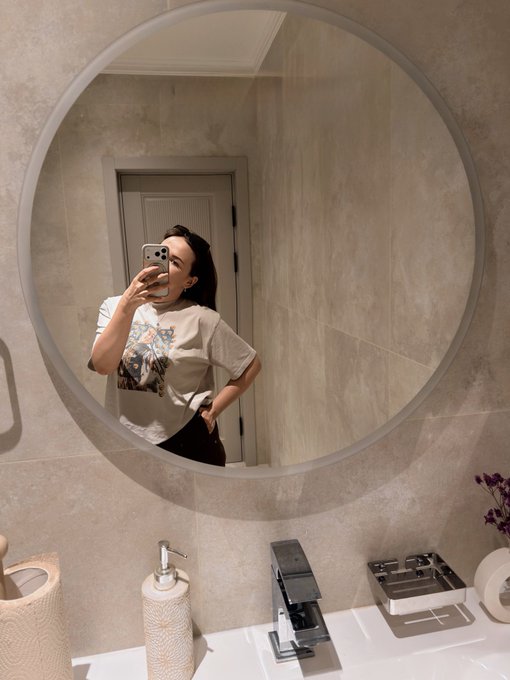 A person takes a mirror selfie in a bathroom, reflected in a large round mirror above a counter with a soap dispenser and small dish.