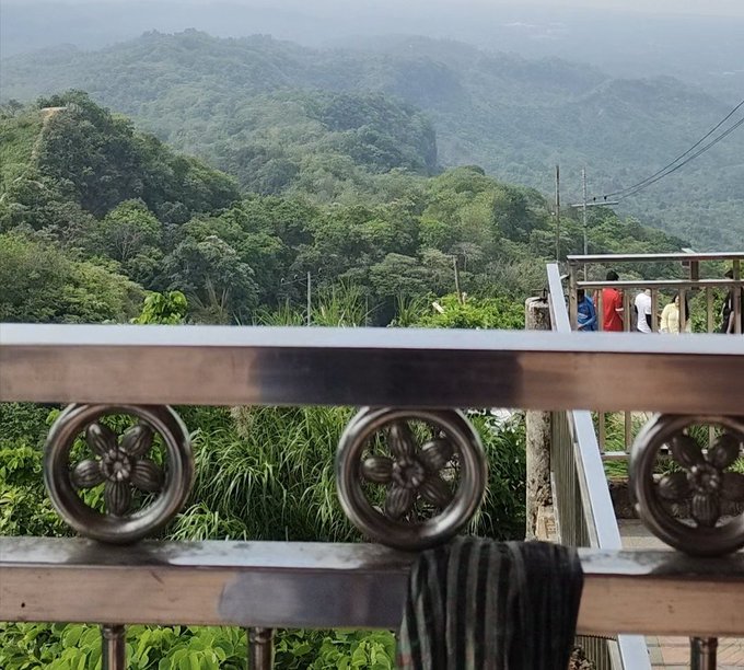 The image shows a metal railing with three circular designs on it, possibly flowers or other decorative elements. The background features a view of mountains and trees, suggesting that this scene takes place in a mountainous area. There are also some clothes hanging from the railing, adding an element of everyday life to the landscape.