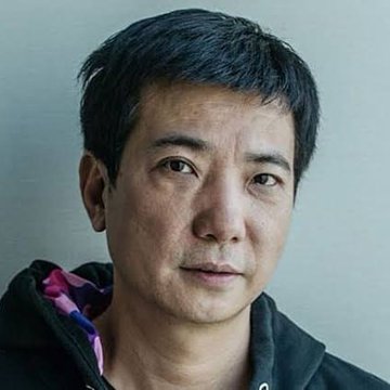 A close-up portrait of an East Asian man with short dark hair, wearing a black hoodie with a pink and purple patterned lining, against a plain gray background.