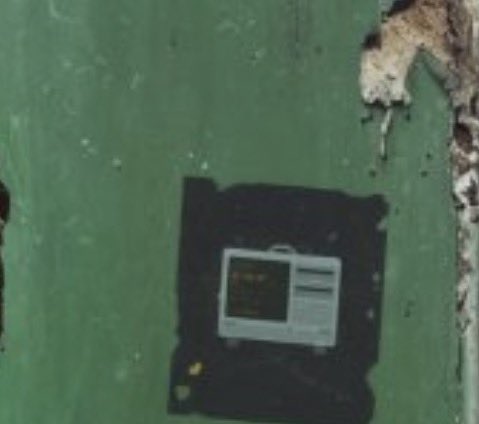 A satellite/aerial view showing what appears to be a damaged building with debris scattered around a green-colored area, possibly near water.