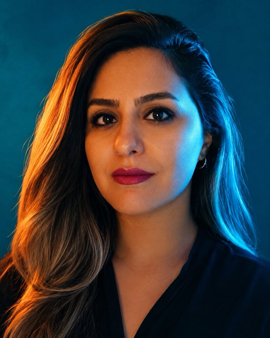 A portrait of a woman with long brown hair, wearing dark lipstick and a black top, illuminated by warm and cool lighting against a dark blue background.