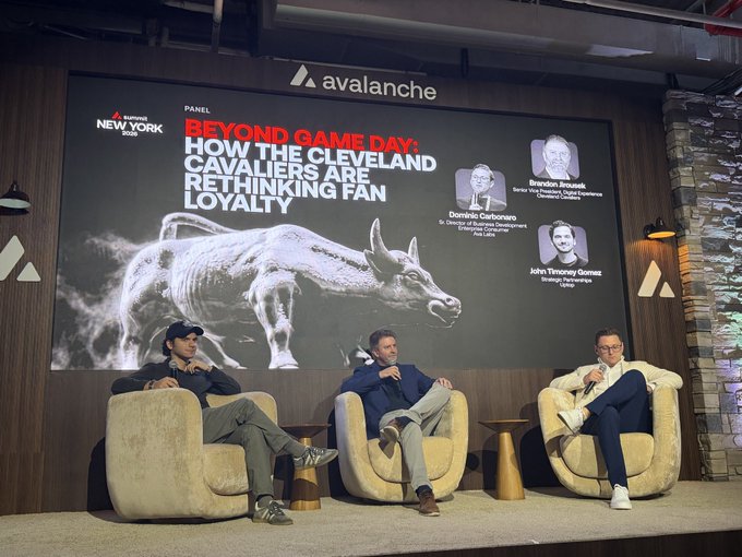 Three men sit on beige chairs on a stage during a panel discussion titled 'BEYOND GAME DAY: HOW THE CLEVELAND CAVALIERS ARE RETHINKING FAN LOYALTY', hosted by Sports Illustrated New York and Avalanche, featuring speakers Brandon Beall, Dominic Castrovince, and John Timoney Gomes.