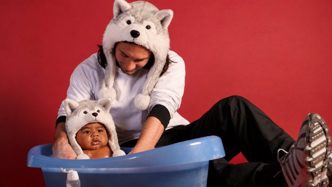 A man and a baby, both wearing matching gray wolf-themed winter hats, pose in front of a red backdrop while the baby sits in a blue plastic tub.