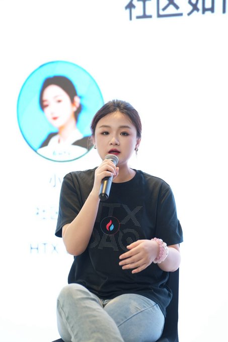 A young woman speaks into a microphone while seated, wearing a black t-shirt featuring the HTX exchange logo, with a backdrop displaying Chinese characters and her own portrait.