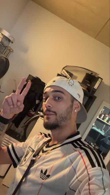 Selfie of a young man wearing a white cap with a jeweled design, an Adidas shirt, and gold chains, making a peace sign indoors.