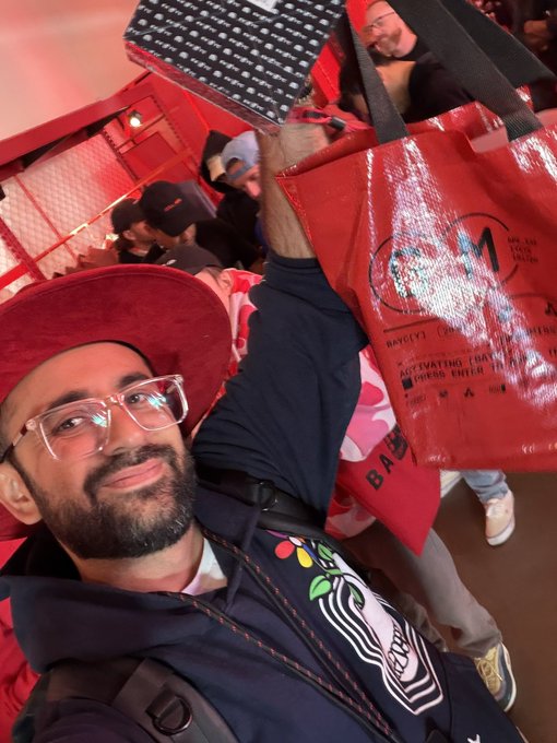 A bearded man wearing a red cap, clear glasses, and a dark hoodie smiles for a selfie while holding up a red tote bag at a crowded event.