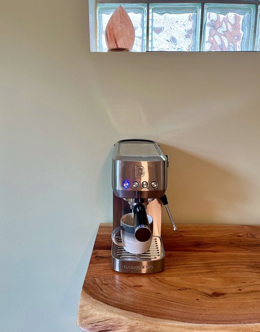 A Casabrews espresso machine on a wooden countertop with a cup placed under the spout beneath a decorative glass block window.