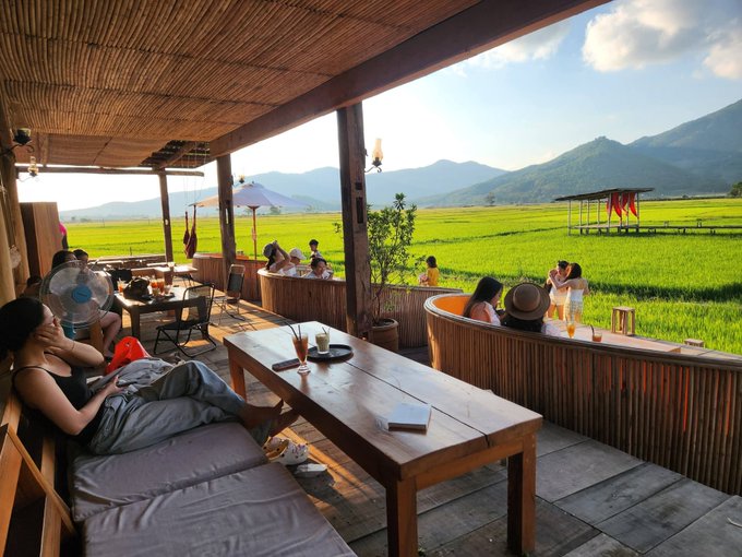 A relaxed open-air cafe with a thatched bamboo roof overlooks lush green rice paddies and mountains, where patrons lounge on cushioned seating and wooden tables.