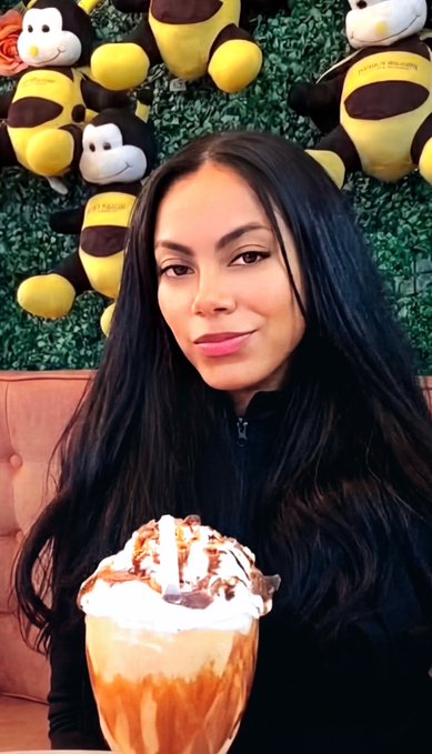 A woman with long dark hair and pink lipstick poses with a large caramel and chocolate-drizzled dessert, seated in front of a green wall decorated with plush toys.