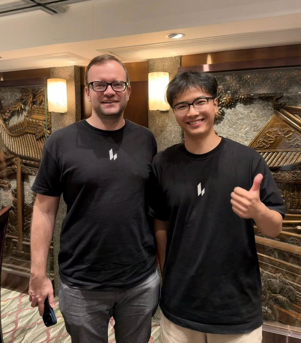 Two men wearing matching black t-shirts with a white logo pose together indoors, with the man on the right giving a thumbs-up.