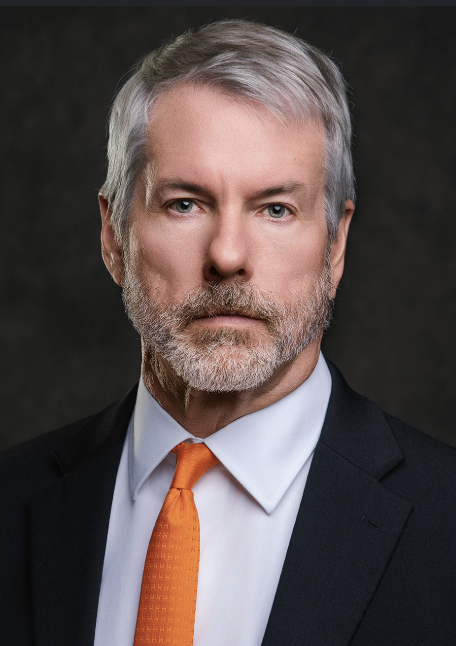 Professional headshot portrait of a distinguished older man with gray hair and beard, wearing a dark suit, white shirt, and bright orange tie against a dark background.
