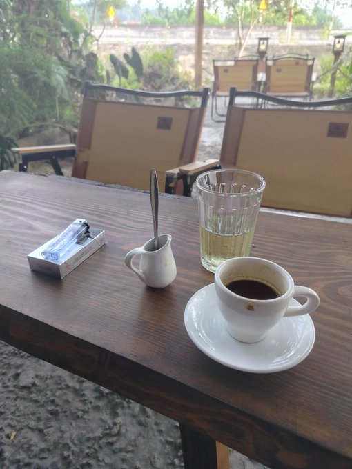 A wooden table at an outdoor café holds a cup of black coffee, a glass of water, and a small pitcher with a spoon, with a pack of "LA Lights" cigarettes nearby. The tranquil scene reflects a quiet moment of reflection, contrasting with the post's eager anticipation of an upcoming market uptrend—suggesting a pause to enjoy a simple pleasure before diving back into the crypto hunt.