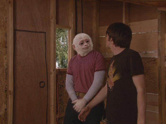 A scene from a film or TV show featuring a person wearing a white monkey mask and a red t-shirt standing in an unfinished wooden room, facing a young man in a black graphic t-shirt.