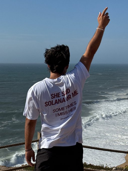 A man stands with his back to the camera, pointing toward the ocean, wearing a white t-shirt that reads 'SHE SAID SOLANA OR ME SOMETIMES I MISS HER'.