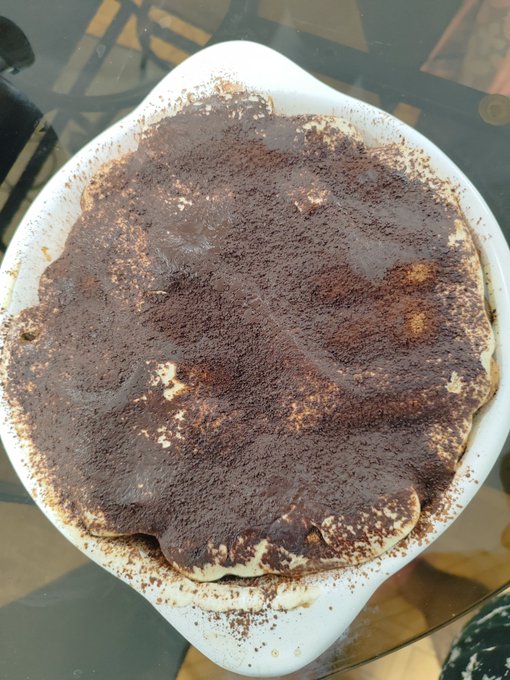 A heart-shaped tiramisu dessert in a white dish, generously dusted with cocoa powder, sitting on a glass table.