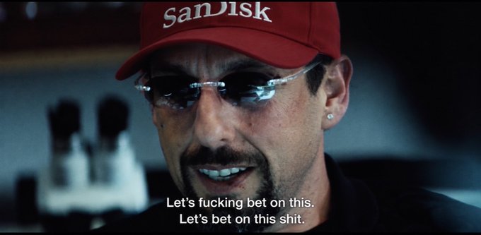 A man wearing a red SanDisk cap and sunglasses in a dark setting with English subtitles reading 'Let's fucking bet on this. Let's bet on this shit.'