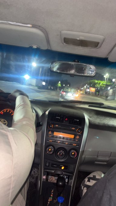 An interior shot from the backseat of a car at night, showing the dashboard with a hand resting on the steering wheel and streetlights visible through the windshield.
