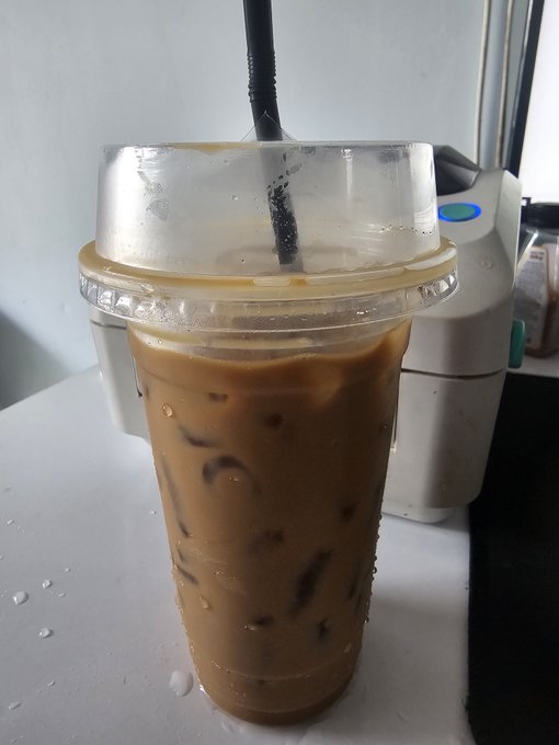 A plastic cup of iced coffee with a black straw sitting on a white surface next to a small appliance.