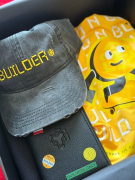 The image features two items - a hat with "builder" written on it in yellow letters and a wallet with stickers for "Builders Only". The hat appears to be made of black fabric, while the wallet has a black leather exterior. Both items are placed inside a box or container, which is sitting on top of a red surface.