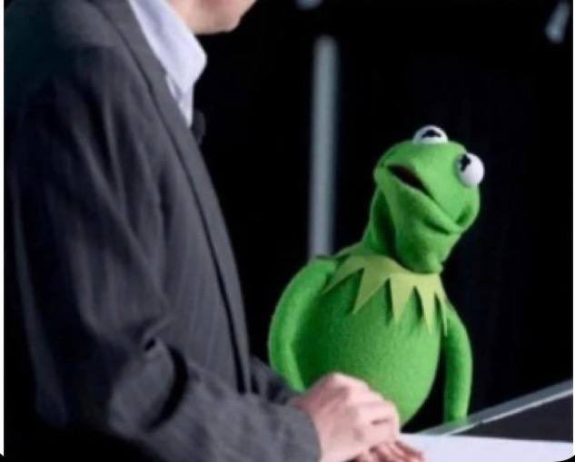Kermit the Frog stands next to a man in a dark suit, looking up at him.