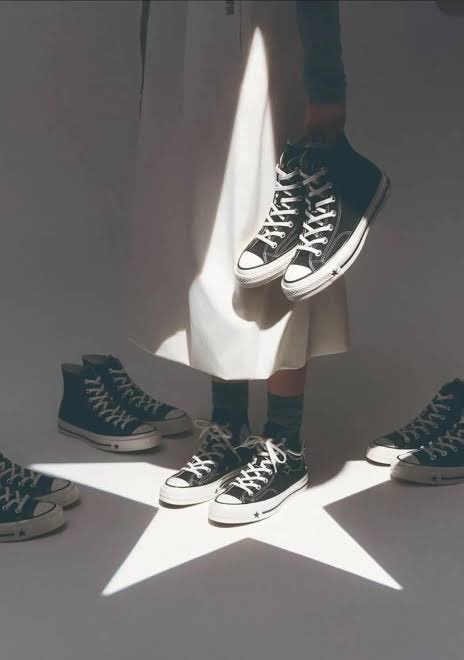 A surreal composition shows a person in a white skirt holding up a pair of black Converse sneakers, while other identical sneakers stand on the floor around a star-shaped light projection.