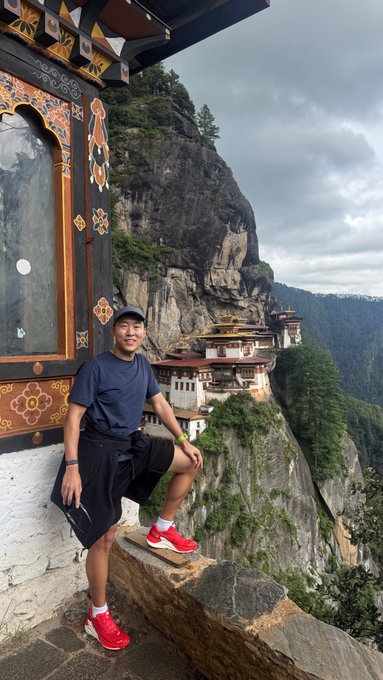 A young man in a blue shirt and red sneakers poses on stone steps outside a traditional Bhutanese monastery perched dramatically on a cliffside, with the Paro Taktsang (Tiger's Nest) visible in the background—capturing the breathtaking setting of the YZi Labs summit in Bhutan mentioned in the post.
