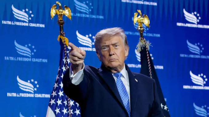 We need to write a concise 1-2 sentence caption describing what is in the image, include any key text, numbers, tickers or names visible. Also note how the image relates to the post. The post is about Trump's crypto earnings, mentioning "特朗普迷因币" (Trump meme coin) and "World Liberty Financial". The image shows Donald Trump pointing, with background "FAITH & FREEDOM COALITION" and flags. So caption: "Donald Trump, wearing a suit, points forward in front of a blue backdrop emblazoned with the 'FAITH & FREEDOM COALITION' logo and two gold‑tipped American flags." That's 1 sentence. Could add that the backdrop also shows "FAITH & FREEDOM COALITION" repeated. That's enough. The relation: It shows Trump, the subject of the crypto earnings claim. So maybe include that the image depicts Trump, linking