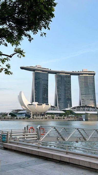A photograph captures the Marina Bay Sands resort in Singapore, featuring its iconic three-tower hotel topped with the SkyPark and the ArtScience Museum shaped like a lotus flower in the foreground, viewed across the water.