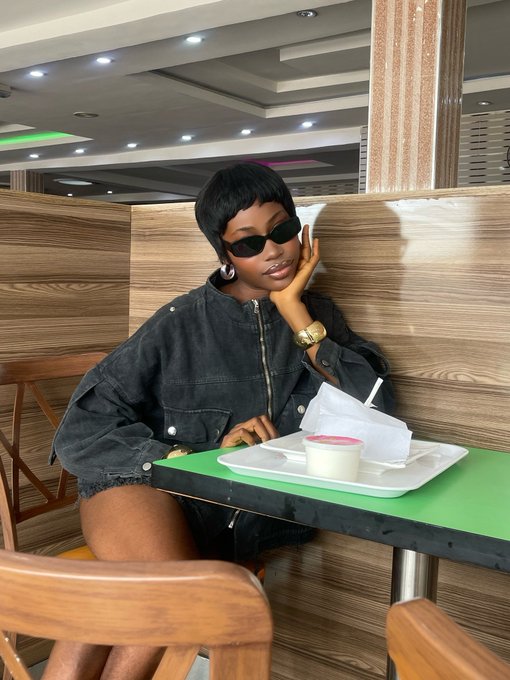 A woman with short black hair, wearing dark sunglasses and a black jacket, poses with her hand on her cheek while sitting at a green table with a food tray.