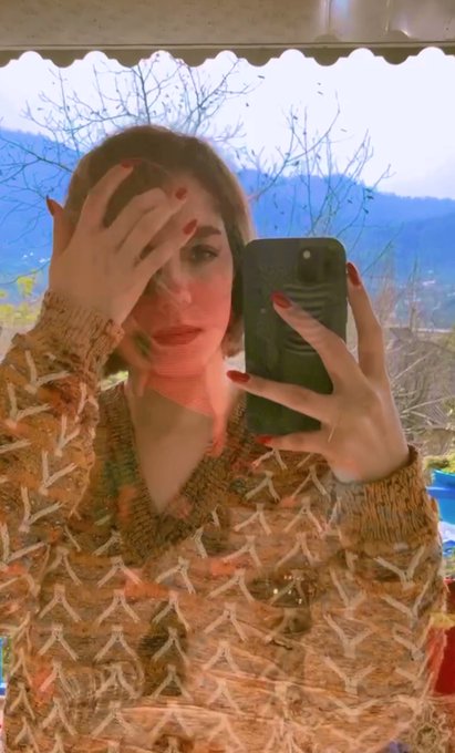 A selfie of a young woman with brown hair taking a photo in a mirror, wearing a textured cream/beige knit sweater, with red nail polish and a dark phone case; a mountainous landscape with bare trees is visible in the background.