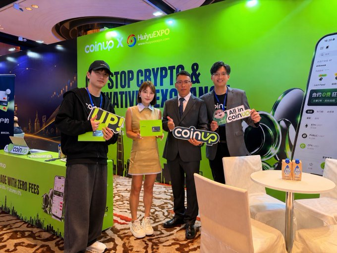 Four people posing at a CoinUp x HuiyuExpo crypto event booth with green branding and the text 'STOP CRYPTO &' partially visible.