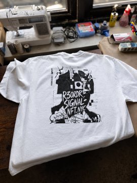 A white t-shirt featuring black graphic artwork with the text "R3ORDR SIGNAL NFT_NYC" is displayed on a desk near a sewing machine and various craft supplies. The shirt represents a small edition release for the NFT NYC convention, showcasing the brand's official merchandise line.