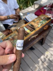 A hand holds a cigar with a 'H.UPMANN' band, with a wooden cigar box and a person visible in the background.
