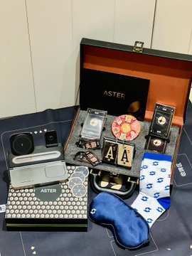 A SafePal and Aster co-branded gift box is displayed open on a dark surface, showcasing its contents including a charger, sunglasses case, eye mask, socks with logos, playing cards, an "ASTER" branded item, and an orange-lined case. This visual aligns with the post celebrating the surprising gift haul and announcing Aster's Mini App integration with SafePal Wallet for on-chain perpetual contracts.