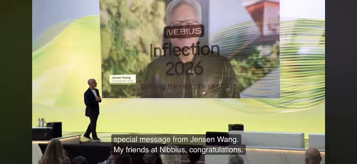 The image shows a stage with a speaker standing behind a podium, giving a speech or presentation. The background of the image features a large screen displaying an image of a man's face along with text that reads "Inflection 2026" and "My friends at nebius, congratulations." This suggests that the event is related to the Inflection 2026 conference held by nebiusai.