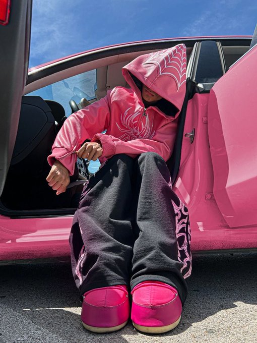 A person wearing a pink hoodie with a spiderweb design, black pants with pink graphics, and bright pink platform shoes sits in the open door of a pink car.