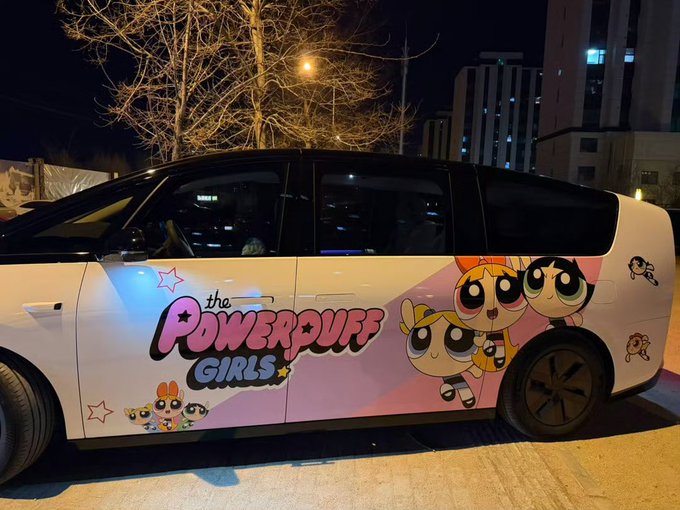 A white MPV is decorated with vibrant "The Powerpuff Girls" graphics at night, featuring the iconic Blossom, Bubbles, and Buttercup characters alongside the show's logo. The playful, eye-catching design makes the van stand out from ordinary vehicles, perfectly matching the post's theme of finding the most stylish and unique ride among countless cars on the street.