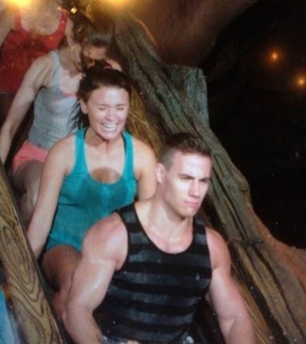 A muscular man in a black tank top sits in front with a serious expression, while a woman in a green tank top rides a wooden water slide behind him, smiling.