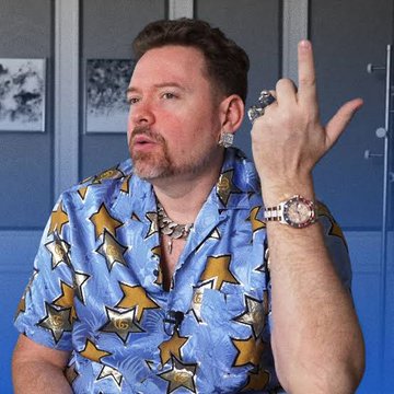 A man wearing a blue shirt with yellow star patterns raises his middle finger while displaying a large wristwatch.