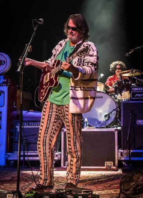 A musician wearing sunglasses and a distinctive striped suit performs on stage with a red guitar, with a drummer visible in the background.