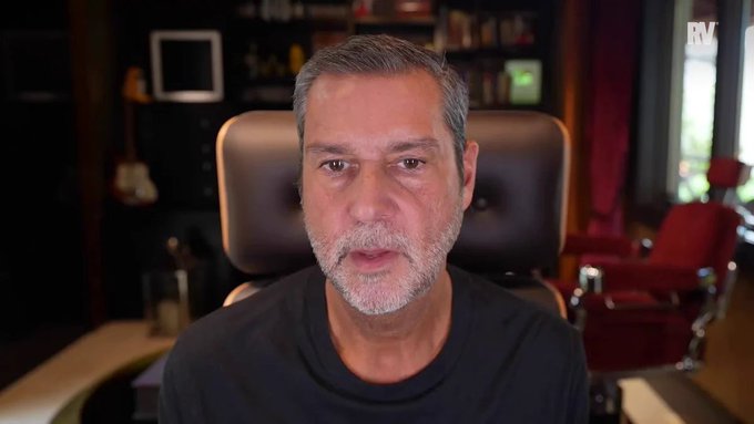 A man with a gray beard speaks directly to the camera from a brown leather chair, with bookshelves, a guitar, and a red chair visible in the background.