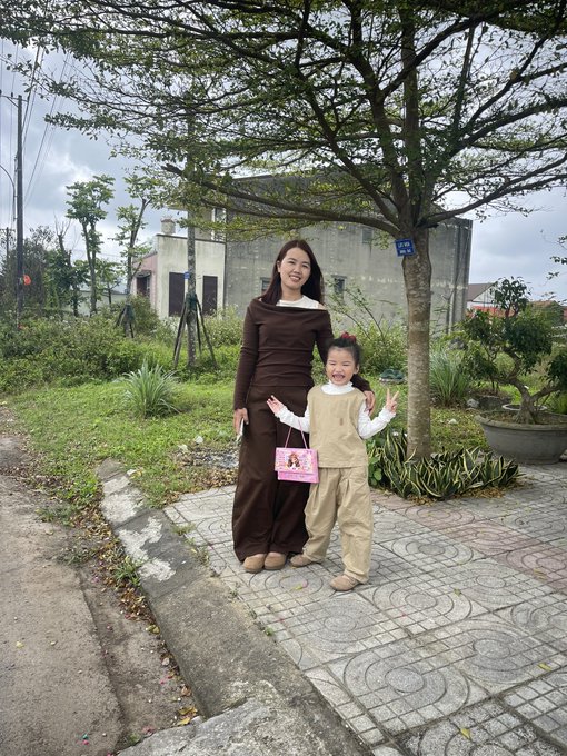 A woman in a brown dress and a young girl in a beige outfit pose smiling on a tiled pathway, both flashing peace signs while the girl holds a small pink gift bag. The cheerful, family-friendly moment aligns with the post's uplifting message of perseverance and starting the day with bright smiles on the "Build X" journey.