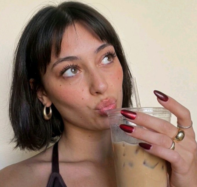 A close-up portrait of a woman with short dark hair, wearing hoop earrings and rings, sipping an iced coffee drink through a straw.