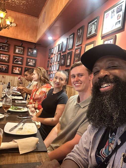 A group of four people smiling for a selfie at a restaurant, with one man in a black cap and a woman in a red dress among them.