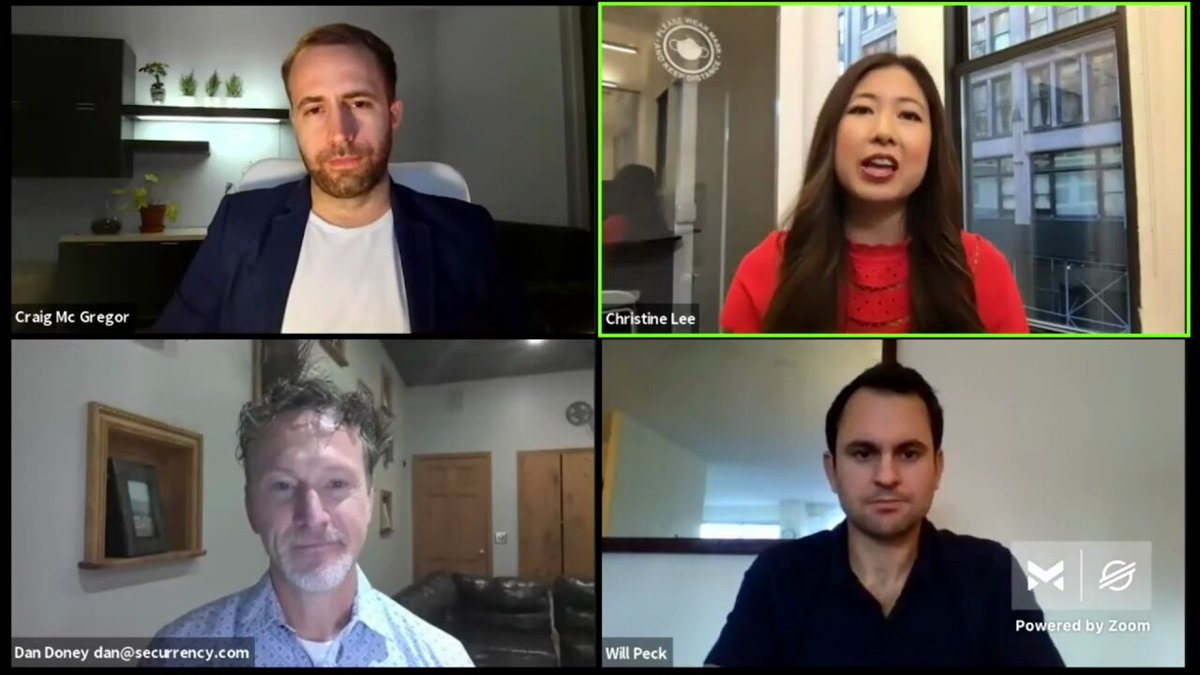 The image shows a group photo of four people sitting around a table for a Zoom call, all looking at the camera. The background reveals that they are in an office setting with cubicles visible behind them. The caption of the image reads "Four men sit together on a video call, with one man speaking into a microphone and another holding up his phone to take a picture."