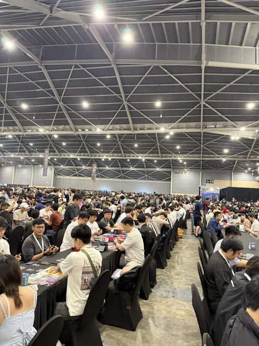 The image shows a massive convention hall filled with rows of tables where crowds are seated playing card games. This relates to the post's lament about missing a 30x gain on $AMC and $MEME calls because the author left their phone behind to attend a card game event like this one, humorously suggesting their priorities lead to "ngmi" status.