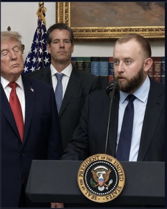 A man speaks at a podium bearing the Seal of the President of the United States, flanked by Donald Trump and another man in a suit, with an American flag in the background.