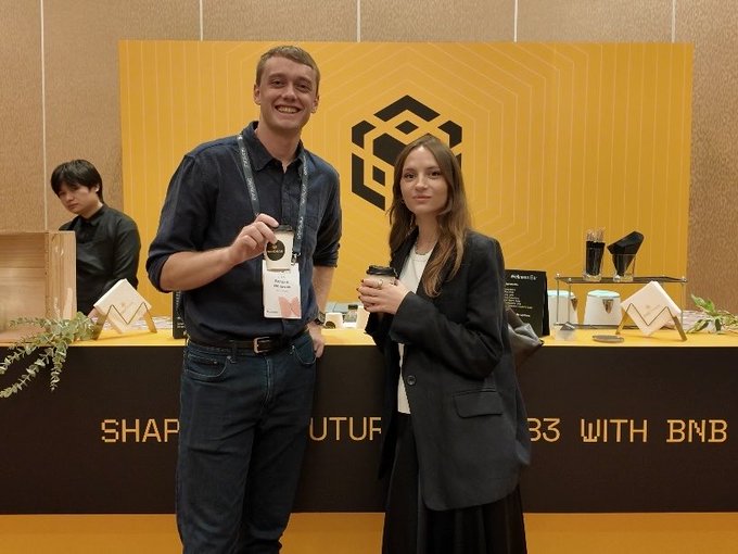 A smiling man and woman holding coffee cups at a Binance booth with a yellow backdrop displaying the BNB logo, and signage mentioning 'BNB' at the counter.